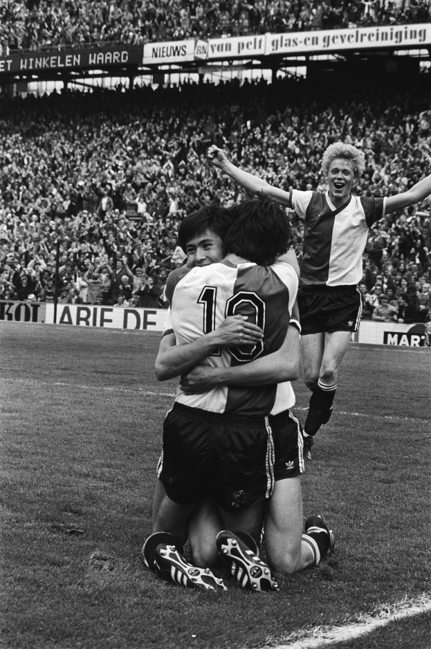 Drei Fussballspieler umarmen sich auf dem Boden beim Feiern eines Tors, mit einer jubelnden Menge und schwenkenden Fahnen im Hintergrund, dargestellt in Schwarz-Weiss.