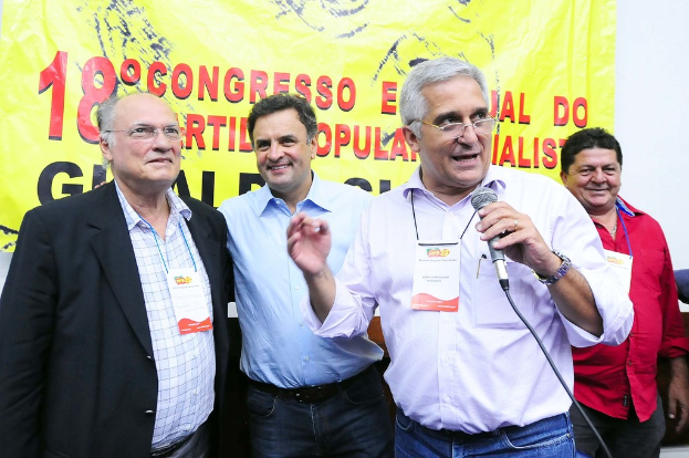 Ein Mann spricht in ein Mikrofon vor einem gelben Vorhang mit der Aufschrift "18 Grad Kongress O", während andere Männer im Hintergrund stehen.