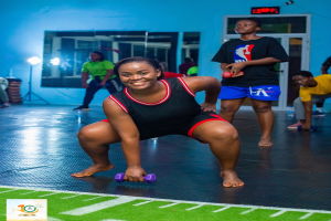 Eine Frau in Kniebeugeposition mit Hanteln in einem Fitnessstudio mit Equipment und Branding im Hintergrund.