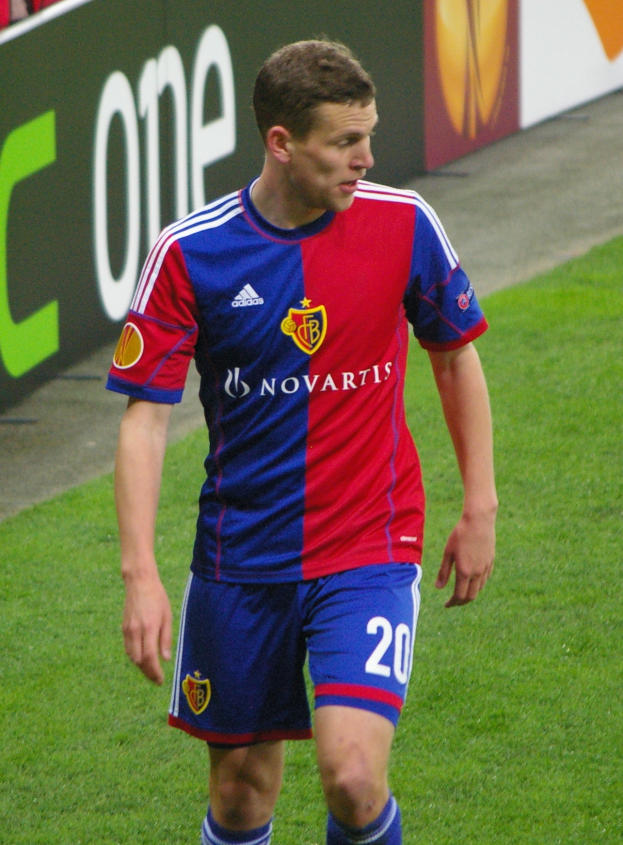 Ein Mann in einem roten und blauen Fußballtrikot, mit ausgestreckten Armen und entschlossener Miene, läuft auf einem Fußballfeld mit einer Tafel im Hintergrund und einigen Menschen auf den Rängen.