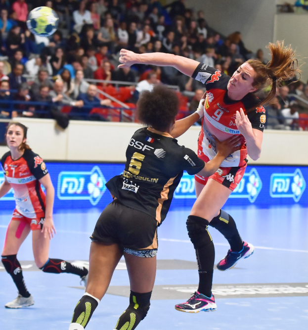 Eine Gruppe von Frauen spielt Handball auf einem Court mit einem Ball in der Luft, beobachtet von Zuschauern in einer Stadionatmosphäre.