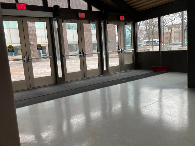 Großer Raum mit hoher Decke, mehreren Fenstern und Türen, mit einem Besen auf dem Boden, informativen Schildern, einem sichtbaren Dach, umliegenden Bäumen, geparkten Fahrzeugen, benachbarten Gebäuden und einem bewölkten Himmel, was auf einen gewerblichen Bodenreinigungsservice hinweist.