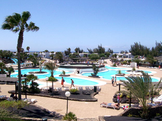Ein Resort mit Pools umgeben von Bäumen, Menschen zwischen ihnen spazieren, Gebäude im Hintergrund und ein klarer Himmel darüber.