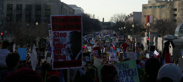 Menschen mit Plakaten auf einer Straße, mit Gebäuden, Bäumen, Laternen und Himmel im Hintergrund.