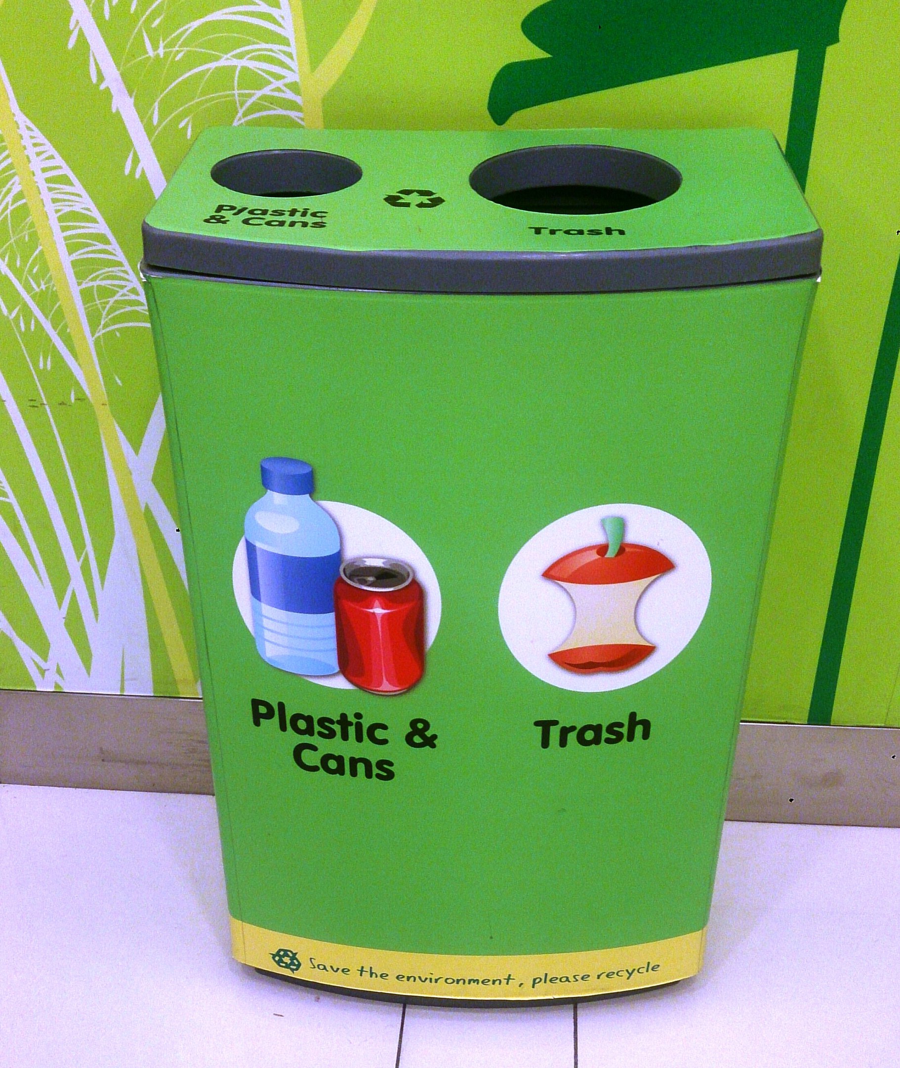 Ein grüner Müllcontainer voller Plastik und Dosen auf einem weißen Boden, mit einem Plakat an der Wand im Hintergrund.