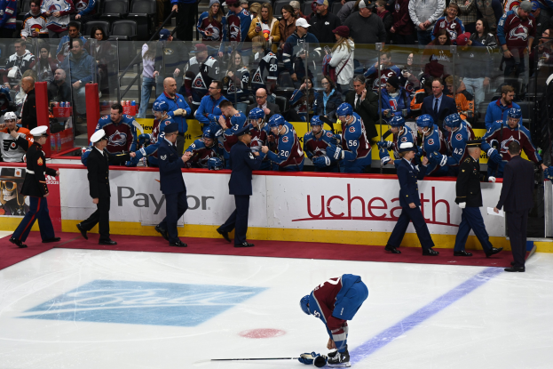 Ein Hockey-Spieler auf dem Eis umgeben von Fans und Team-Mitarbeitern, mit Zuschauern hinter den Boards mit der Aufschrift "Colorado Avalanche vs San Jose Sharks - 3/24/15 NHL Pick, Odds, and Prediction."