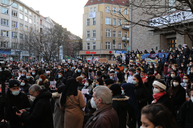 Eine große Gruppe von Menschen mit Mund-Nasen-Schutz steht vor einem Gebäude mit Fenstern, Bäumen, Schildern und Ampeln unter einem bewölkten Himmel, einige halten Schilder und Mobiltelefone, was auf eine Protestaktion gegen ein Regierungsverbot hinweist.