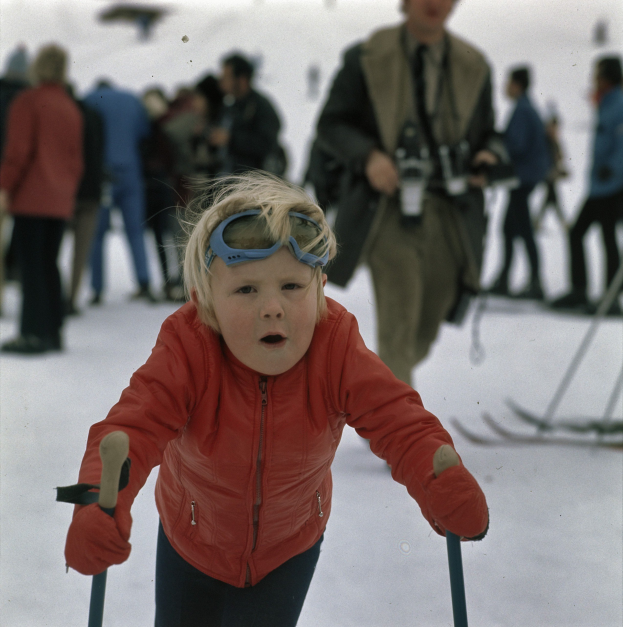 Ein junger Junge in roter Jacke und Skibrille fährt eine schneebedeckte Anhöhe mit Skistöcken hinunter, umgeben von mehreren Menschen und einigen mit Snowboards im Hintergrund.