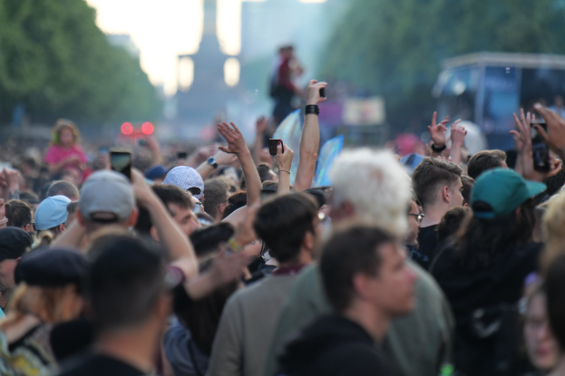 Eine Menge bei einem Musikfestival mit erhobenen Händen, einige tragen Mützen und halten Telefone, vor einem unscharfen Hintergrund aus Bäumen, Gebäuden, Lichtern und einem Fahrzeug.