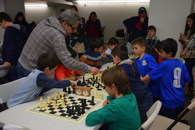 Children are playing chess in a competition while adults watch from the background and sides.