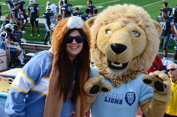 Eine Frau steht in der Nähe einer Person in einem Löwenkostüm, mit Spielern, die hinter ihr auf dem Boden stehen und Zuschauern, die auf Bänken und Stühlen links sitzen.