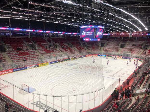 Ein Hockey-Spiel in einer großen Arena mit Zuschauern auf den Sitzplätzen und Stehplätzen, einem Zaun um die Eisfläche, einem Bildschirm mit Text und Metallrahmen sowie einer Decke mit Deckenbeleuchtung.