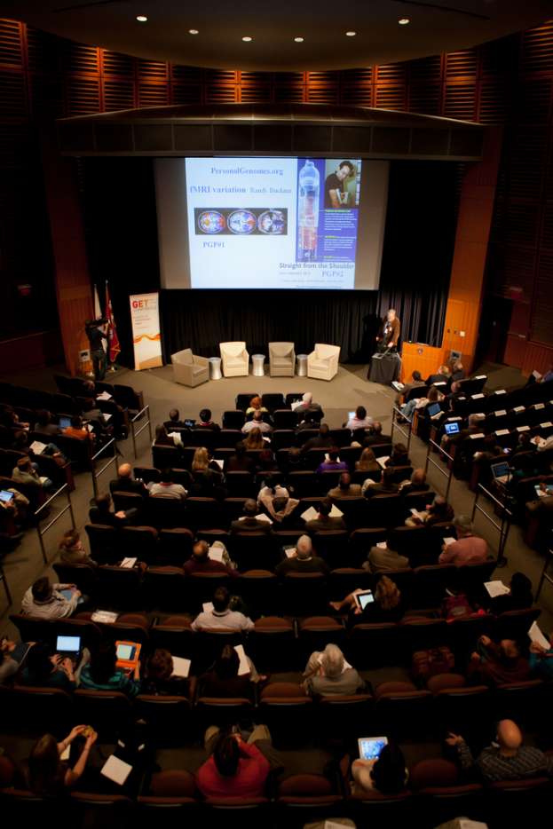 Ein mit sitzenden Menschen gefüllter Saal, beleuchtet von vielen Lichtern, mit Fahnen links, einem Podium rechts und einer Werbetafel links.