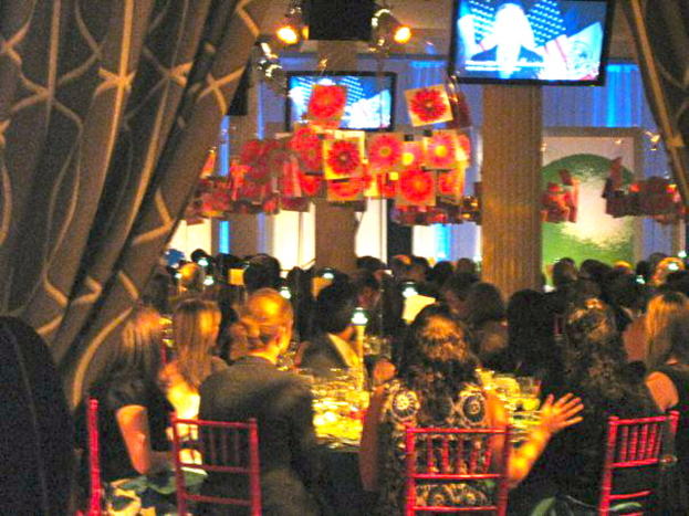 Eine Gruppe von Menschen sitzt an Tischen mit Essen, Fernsehern, Lampen, hängenden Papieren und Vorhängen in einer scheinbar feierlichen Umgebung.