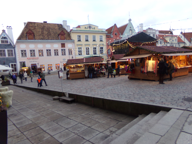 Ein lebendiger Weihnachtsmarkt in Tallinn, Estland, mit Menschen um beleuchtete Stände, festliche Dekoration, Gebäude mit Fenstern im Hintergrund, Treppen, die zum Markt führen, und Töpfe mit Pflanzen, die herumstehen.