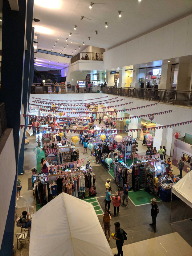 Ein volles Einkaufszentrum mit Ständen, hängenden Kleidern, Dekorationsfahnen, Ballons, sitzenden und stehenden Menschen, Geländern, Säulen und Deckenleuchten, das an einen Weihnachtsmarkt im Mall of Asia erinnert.