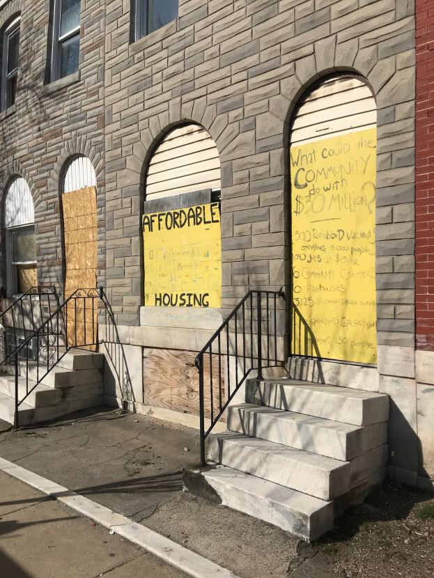 Ein verbarrikadiertes Gebäude mit einer Tafel, die "Bezahlbarer Wohnraum" lautet, eine Treppe mit Geländern, die zum Eingang führt, und eine Straße unten.