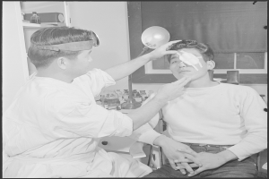 Ein Mann sitzt in einem Stuhl, während ein Arzt seine Augen mit einem tragbaren Instrument untersucht, mit medizinischen Gegenständen auf einem Tisch und einem Vorhangfenster im Hintergrund.