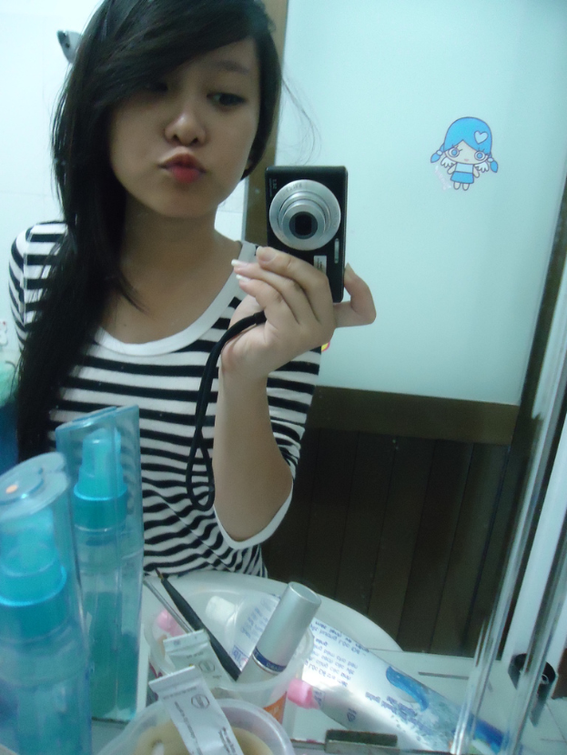 Eine Frau steht vor einem Schminktisch mit Kosmetikartikeln und macht ein Foto mit einer Kamera, wobei im Hintergrund eine Tür zu sehen ist.