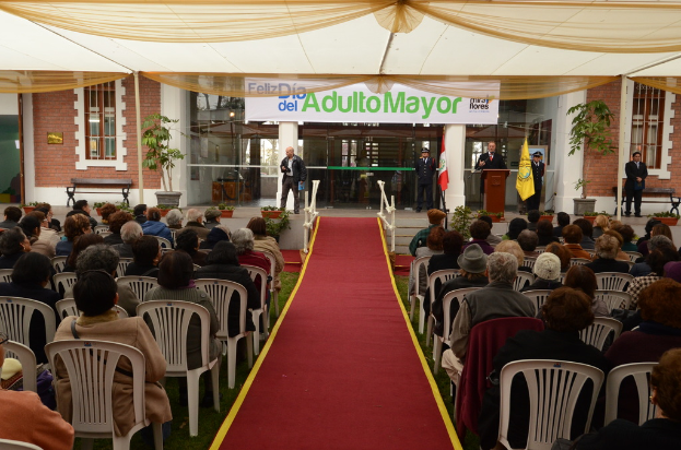 Ein Mann steht vor einem Gebäude mit einem Bogen, auf dem "Adulto Bürgermeister" steht, während viele Menschen im Hintergrund auf Stühlen sitzen.