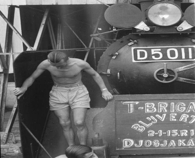 Zwei Männer in Schwarz und Weiß stehen auf einem Zug neben einer Tafel mit Text, mit Metallstangen und einer Wand im Hintergrund.