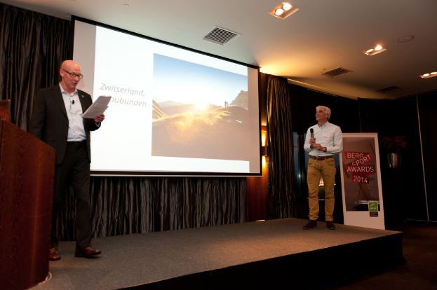 Zwei Männer stehen auf einer Bühne, einer hält ein Mikrofon und der andere hält Papiere, mit einem Rednerpult, Projektionsdisplay, Vorhängen, Banner, Fenster und Bühnenscheinwerfern.
