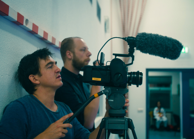 Ein Mann in einem blauen T-Shirt filmt mit einer Kamera, wobei ein Mikrofon darüber zu sehen ist, während ein Mann in einem schwarzen Hemd und eine Frau in einem weißen Kleid anwesend sind, mit einer weißen Wand und einem Vorhang im Hintergrund.