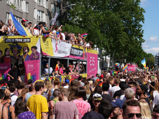 Eine große Menschenmenge marschiert auf der Straße bei der Berlin Pride 2019, viele tragen Mützen und Schutzbrillen, einige halten Fahnen, mit Bannern, Laternenmasten, Bäumen und Gebäuden mit Fenstern im Hintergrund bei bewölktem Himmel.