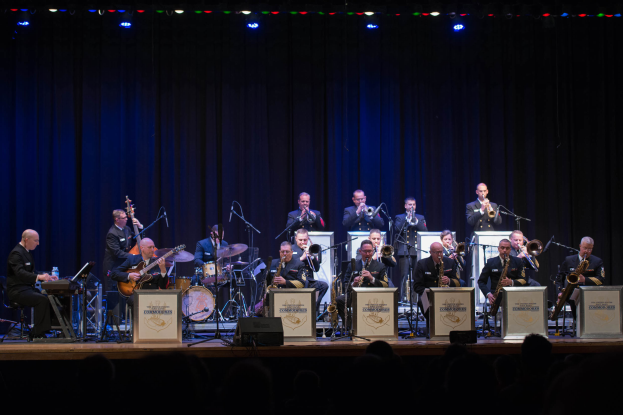 Eine Gruppe von Musikern auf der Bühne, die Instrumente spielen, mit Mikrofonen und Ständern, Schilder mit der Aufschrift "The New Orleans Jazz Band", ein Vorhang im Hintergrund und ein Publikum unten, das von Scheinwerfern beleuchtet wird.