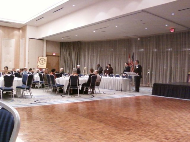 Eine Gruppe von Menschen, die auf Stühlen sitzen und einige stehen, umgeben von Fahnen auf Stangen, Lampen und Schildern, mit Vorhängen im Hintergrund.