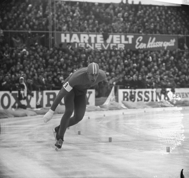 Ein Schwarz-Weiß-Foto eines Eisschnellläufers in Aktion auf dem Eis, der einen Helm und Eislaufschuhe trägt, mit Zuschauern und Tafeln mit Wörtern im Hintergrund.