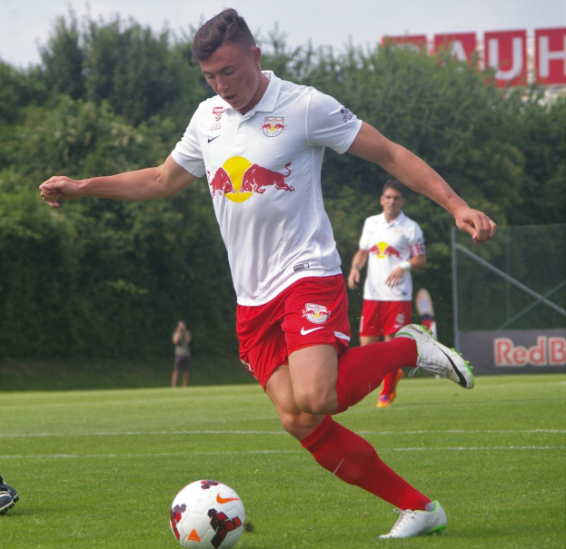 Ein Mann in einem Fussballtrikot kickt einen Ball auf einem grasigen Feld mit Bäumen und einem klaren blauen Himmel im Hintergrund, mit einem Schild mit der Aufschrift "RB Leipzig" sichtbar.