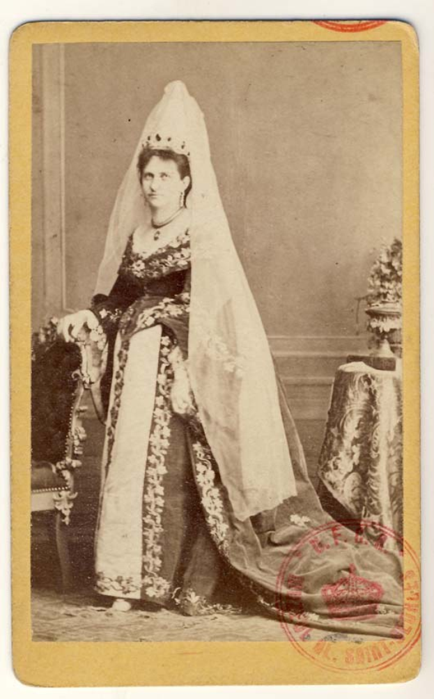 Altes Schwarz-Weiß-Foto einer Frau in traditionellem russischer Hochzeitskleid, das auf einem Stuhl sitzend einen Blumenstrauß hält, mit einem Blumenstrauß auf einem Tisch zu ihrer Rechten und Text plus einem Stempel unten.