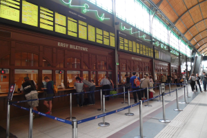 Leute in einer Warteschlange an einem Bahnhofsschalter mit Taschen, Stangen mit Bändern, Informationsschildern, Deckenleuchten und Treppen mit Geländern im Hintergrund.