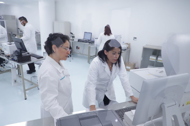 Laboratorszene mit Forschern in weißen Laborkitteln, die an Tischen mit Monitoren, Tastaturen und Deckenbeleuchtung auf Computern arbeiten, mit Schränken und einer Wand im Hintergrund.