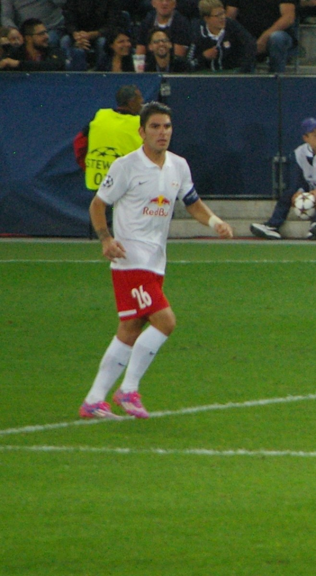 Ein professioneller Fußballspieler in einem weißen Trikot und roten Shorts kickt einen Ball auf einem Feld, mit Zuschauern im Hintergrund.