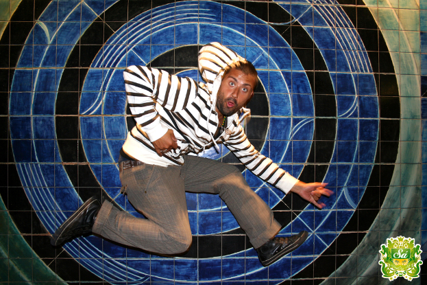 Ein Mann tanzt vor einem bunten Hintergrund mit einem Logo in der rechten oberen Ecke.