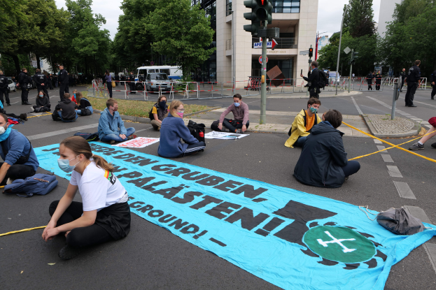 Eine Gruppe maskierter Personen sitzt vor einem Gebäude auf dem Boden und hält ein Transparent mit Text, umgeben von verstreuten Taschen und Gegenständen, mit einer Ampel, Bäumen und anderen Gebäuden im Hintergrund.