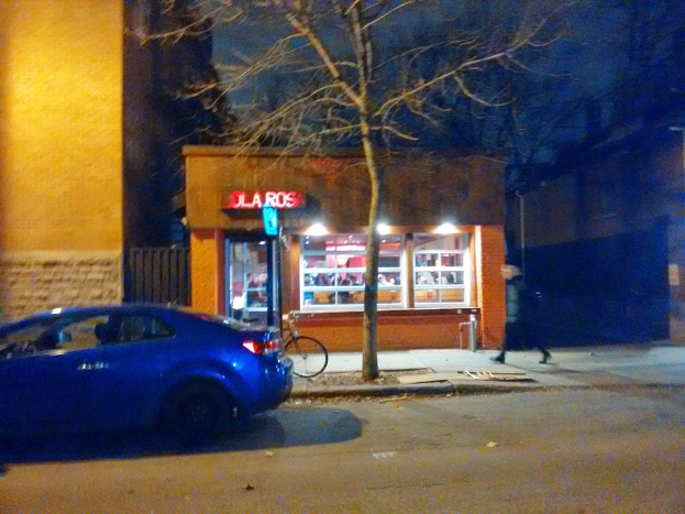 Eine nächtliche Straßenszene mit Fahrzeugen auf der Straße, Fahrrädern neben Pfählen, einer Uhr an einem Pfahl, beleuchteten Gebäuden im Hintergrund und Menschen auf dem linken Gehweg.