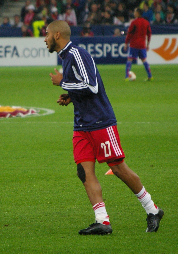 Ein professioneller Fussballspieler in einem blauen Trikot und roten Shorts läuft auf einem Feld, umgeben von sitzenden Zuschauern und Werbetafeln.