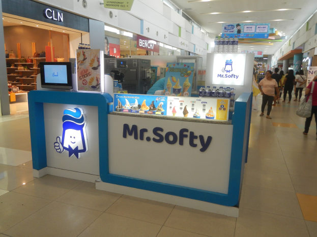 Mr. Softy-Eiscafé-Innenraum in einem Einkaufszentrum mit Theke, Monitor, Menschen und Displayregalen im Hintergrund.