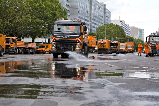 Eine Gruppe von Volvo-Lkw fährt auf einer Straße neben hohen Gebäuden, während Menschen auf der rechten Straßenseite stehen, Bäume und Gebäude im Hintergrund zu sehen sind und der Himmel oben sichtbar ist.