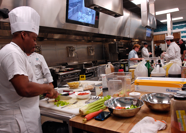 Eine Gruppe von Köchen in einer Küche, mit einem im Vordergrund in einer weißen Schürze und Mütze, der ein Messer hält und von Utensilien, Schalen, Gemüse, einem Handy, Flaschen und anderen Gegenständen umgeben ist, während im Hintergrund ein Fernseher und Deckenlampen zu sehen sind.