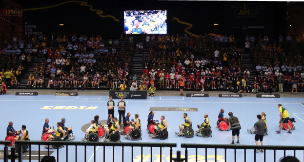 Eine Gruppe von Rollstuhlfahrern auf einem Court bei den Invictus Games Düsseldorf 2020, umgeben von einem Geländer und einer großen Zuschauermenge, mit Stufen zu einem Bildschirm und Informationsplakaten im Hintergrund.
