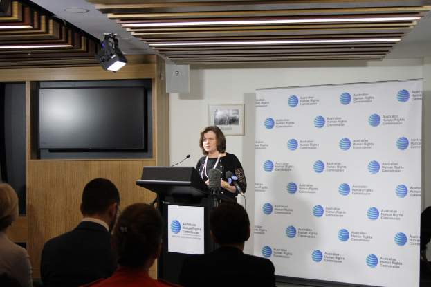 Eine Frau steht auf einer Bühne und spricht in ein Mikrofon, vor ihr sitzen Zuschauer, an der Wand hängt ein Banner, ein Fernseher an der Holzwand und Lampen an der Decke.