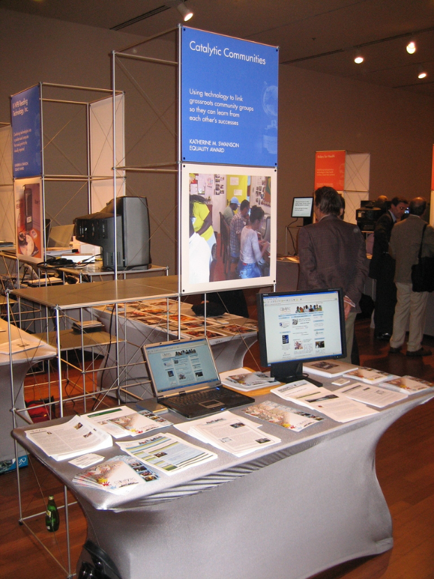 Ein Geschäft mit einem zentralen Tisch, auf dem Bücher, ein Laptop und ein Desktop-Computer liegen, umgeben von Bücherregalen, mit Werbetafeln, einem Fernseher und Deckenleuchten im Hintergrund und ein paar Menschen auf der rechten Seite.