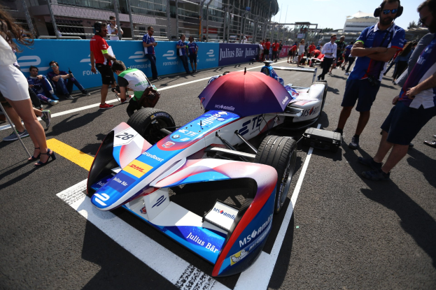 Formula E Auto mit weißer, blauer, roter und schwarzer Lackierung auf einer Rennstrecke umgeben von Menschen, mit einem Schirm im Vordergrund und Rennstrecken-Infrastruktur im Hintergrund unter einem klaren blauen Himmel.