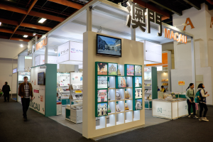 Eine Gruppe von Menschen steht vor einem Messestand auf der Macau International Book Fair, der mit Büchern und anderen Gegenständen gefüllt ist, einem montierten Fernseher, Namensschildern und von Lichtern beleuchtet, wobei einige Besucher Taschen tragen.
