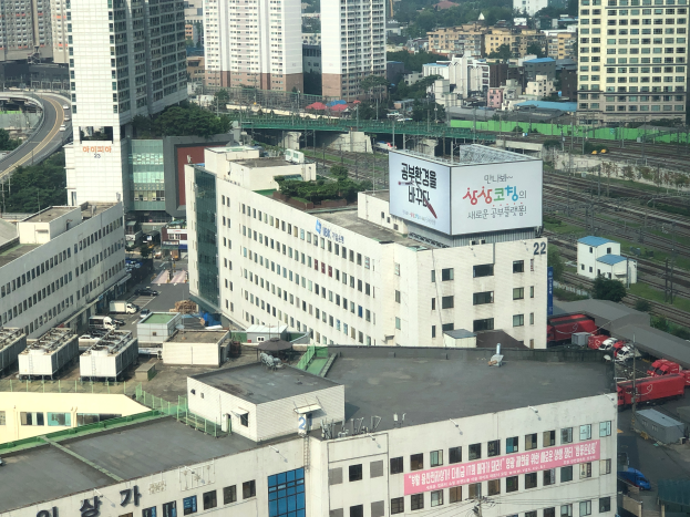 Eine Stadtansicht von einem hohen Standpunkt aus, mit zahlreichen Gebäuden, Straßen mit Fahrzeugen, Bahnschienen, Masten, Bäumen und einer prominenten Brücke im Vordergrund, mit einem Plakat in der Mitte.