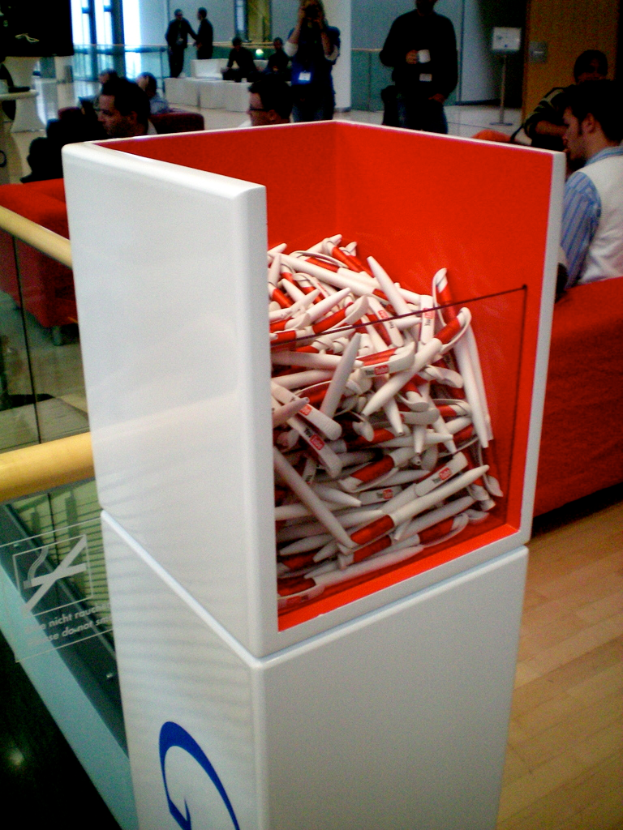 Ein Kasten mit Stiften im Vordergrund, mit Gruppen von Menschen im Hintergrund, einem Schild an einem Pfahl, einer Wand, einem Holzboden und Text auf einem Spiegel.